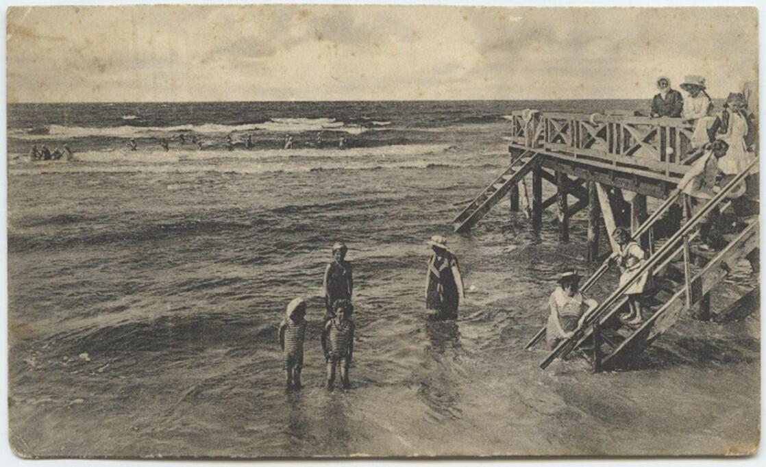 Moterys ir vaikai maudosi prie Palangos jūros tilto, 1905 m. Damos dėvi maudymosi sukneles ir skrybėles, o vaikai – uždarus maudymosi kostiumus. P. Mongirdaitės atvirukas.