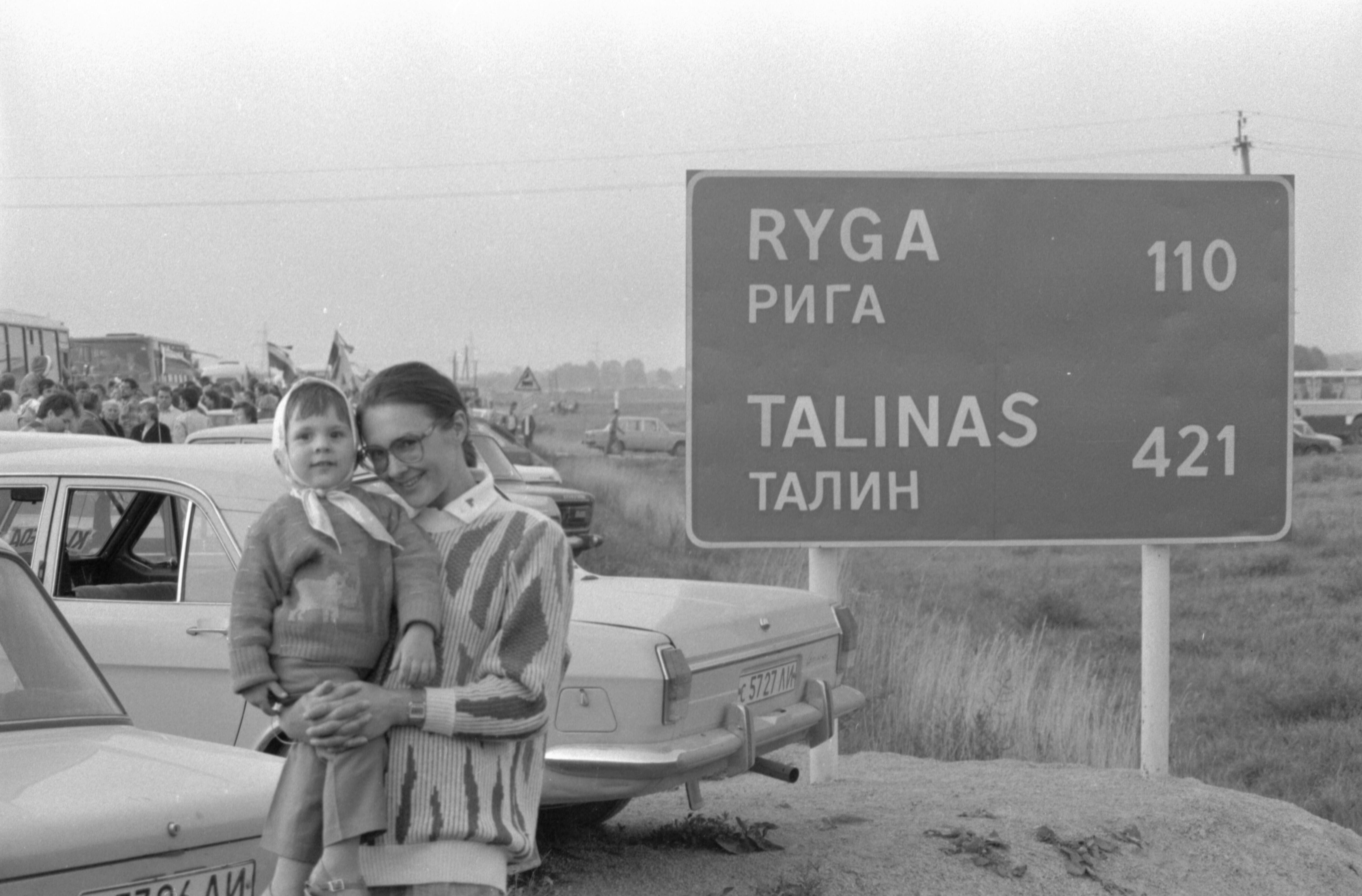 Fotografas, žurnalistas iš Klaipėdos Antanas Stanevičius fotografuoja Baltijos kelio akcijos dalyvius ruože prie Pasvalio. Kelio ženklas, informuojantis apie atstumus iki Rygos ir Talino. Stovi lituanistė Eglė Zalatoriūtė su dukra Elvyra.